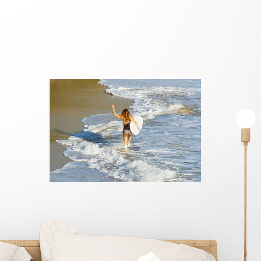 Woman running along beach with surfboard Wall Mural