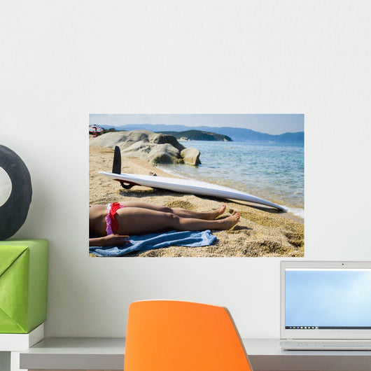 Young woman sunbathing on beach with surf board on sand Wall Mural