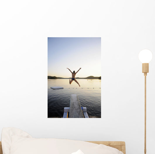 A swimmer jumps off a diving board as the sun sets over a lake Wall Mural
