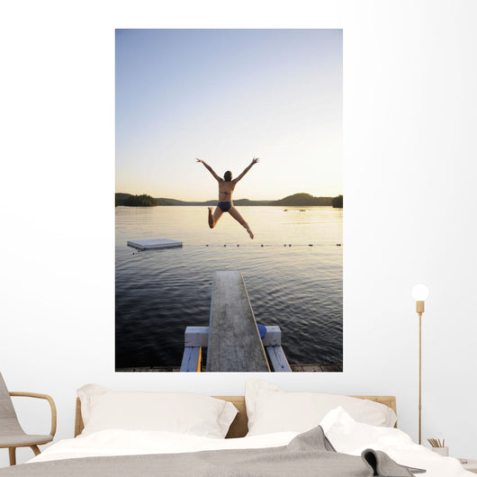 A swimmer jumps off a diving board as the sun sets over a lake Wall Mural