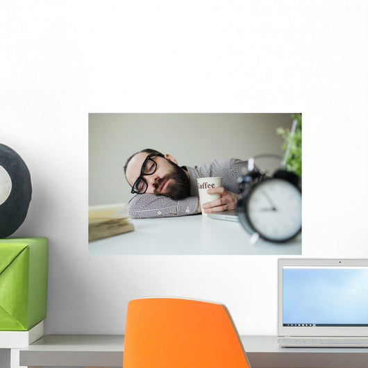 Man sleeps in office on table over laptop with coffee in hand Wall Mural