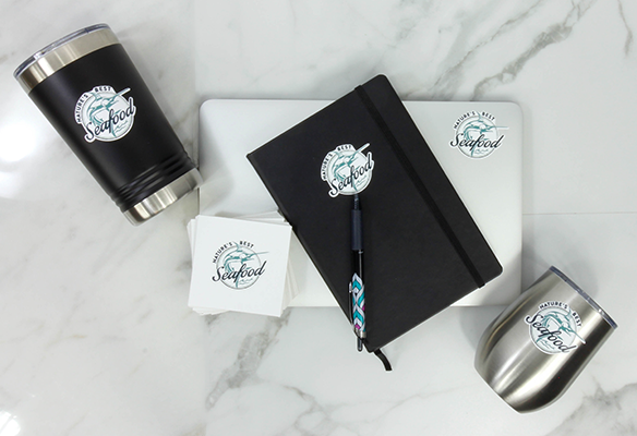 Small vinyl sticker decals showing a local business logo. Stickers are used on a laptop computer, black journal, coaster, and two mugs. 