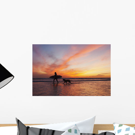 A Surfer Walks In Shallow Water With Her Dog At Sunset Wall Mural