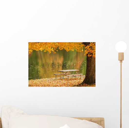 A picnic table beside a tranquil lake with golden leaves on the trees Wall Mural