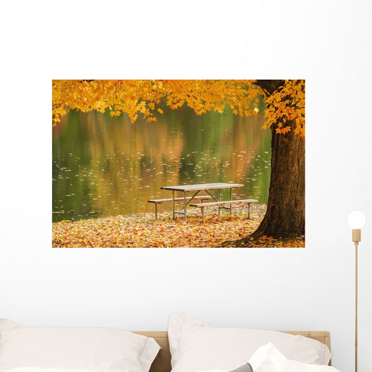 A picnic table beside a tranquil lake with golden leaves on the trees Wall Mural