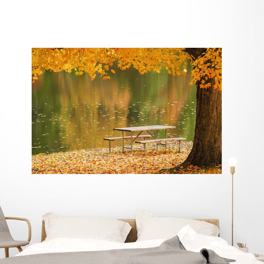A picnic table beside a tranquil lake with golden leaves on the trees Wall Mural