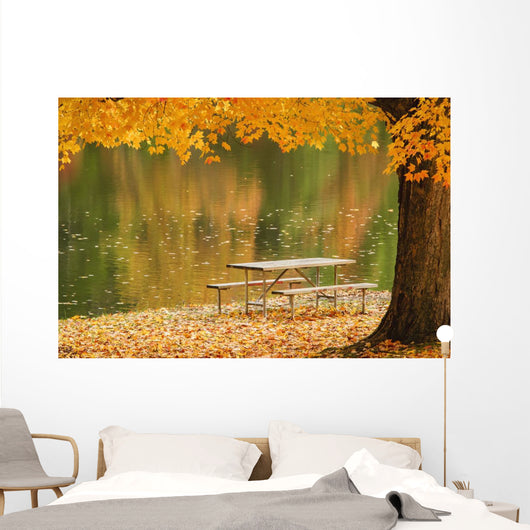 A picnic table beside a tranquil lake with golden leaves on the trees Wall Mural