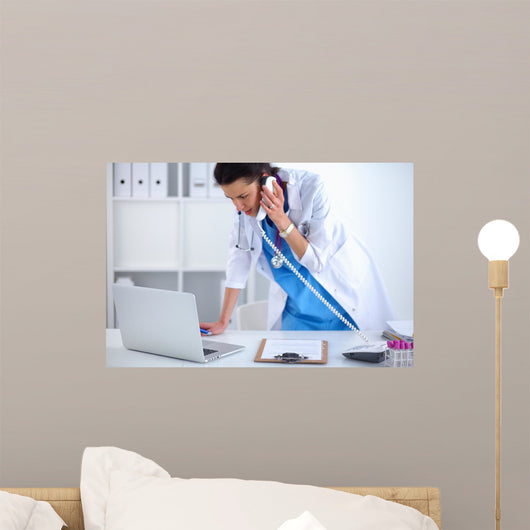 Young woman doctor in white coat at computer using phone Wall Decal