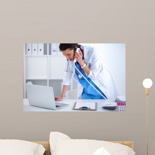 Young woman doctor in white coat at computer using phone Wall Decal