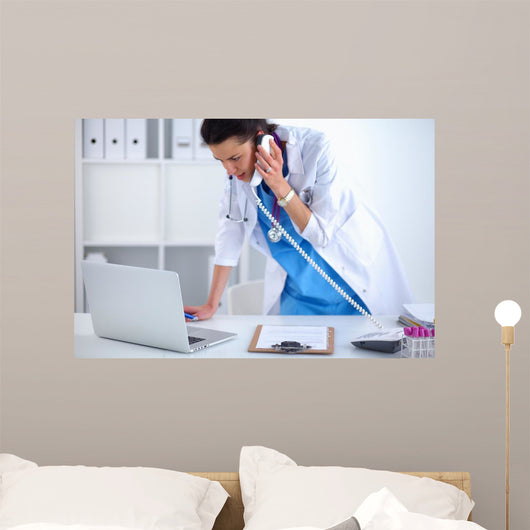 Young woman doctor in white coat at computer using phone Wall Decal