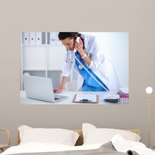 Young woman doctor in white coat at computer using phone Wall Decal
