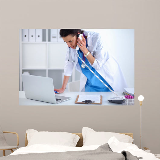 Young woman doctor in white coat at computer using phone Wall Decal