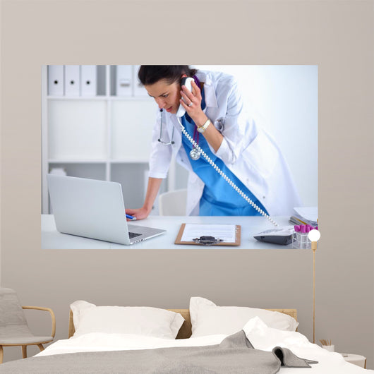 Young woman doctor in white coat at computer using phone Wall Decal